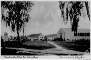 Leegebruch – Wasserwerk, Blick aus nördlicher Richtung auf das fertig gestellte Wasserwerk und die dahinterliegenden Ledigenwohnheime (links im Bild das Haus mit der Nummer 1). Im Vordergrund, linksseitig deutlich zu sehen, eines der fünf „Brunnenhäuser“ (Tiefbrunnen). Aus dem Gesamtbild ist auch zu entnehmen, dass die Ausbildung der Straßenzüge und der Grünflächen dem Gesamtbautempo hinterherlief – unfertige Anlagen.